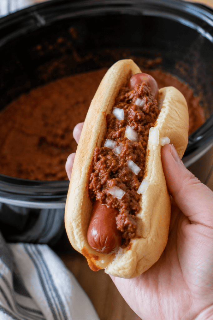 Hand holding a chili dog topped with homemade hot dog chili and diced onions.
