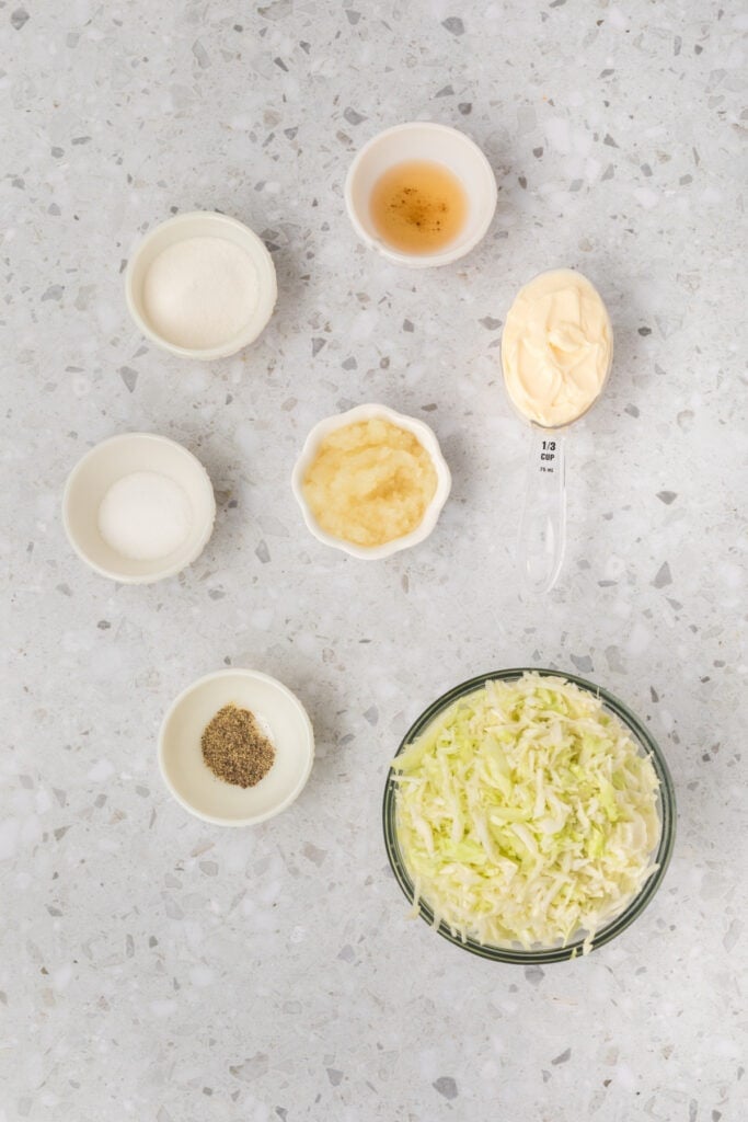 Ingredients for Southern-Style Slaw arranged on a countertop, including shredded cabbage, mayonnaise, grated onion, apple cider vinegar, sugar, salt, and black pepper.
