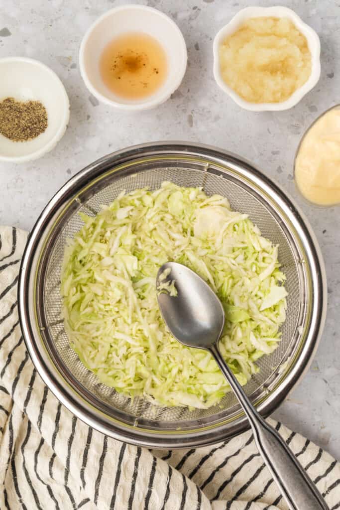 Spoon pressing shredded cabbage in a colander to squeeze out excess liquid.