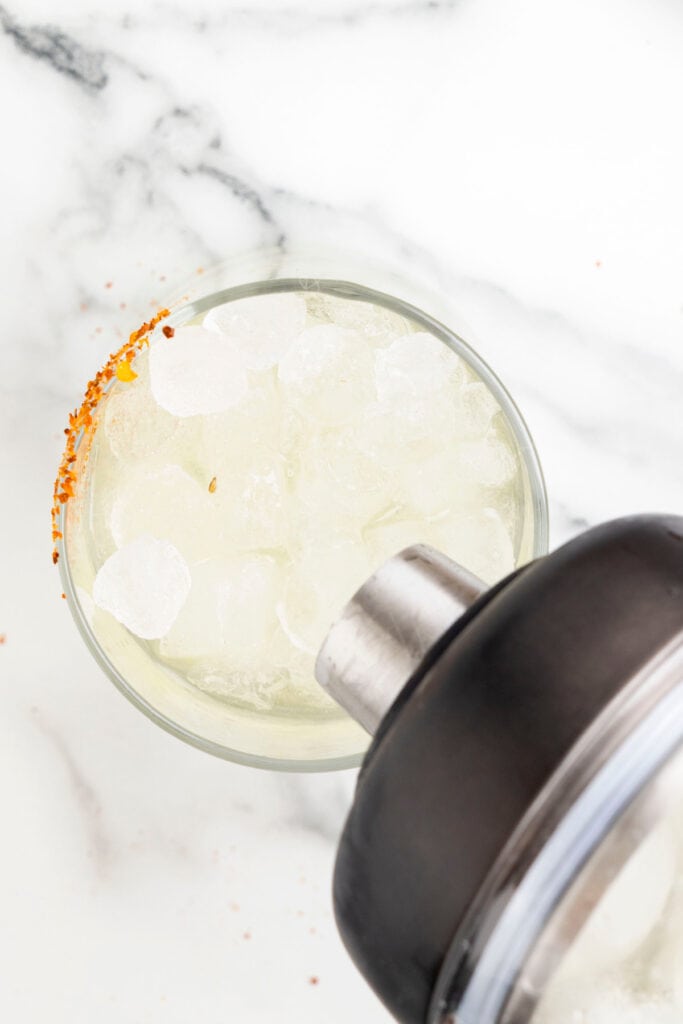 Pouring a freshly shaken spicy margarita into a prepared glass with ice and a Tajín rim