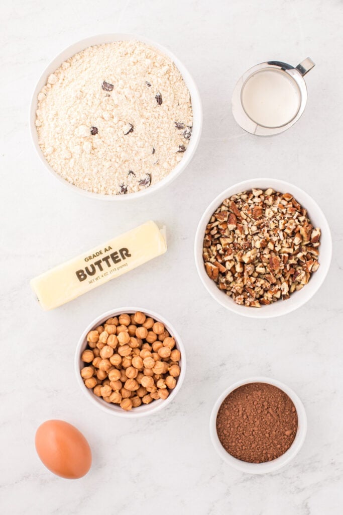 Ingredients for Easy Turtle Cookies including chocolate chip cookie mix, cocoa powder, butter, egg, heavy cream, chopped pecans, and caramel bits arranged on a countertop.