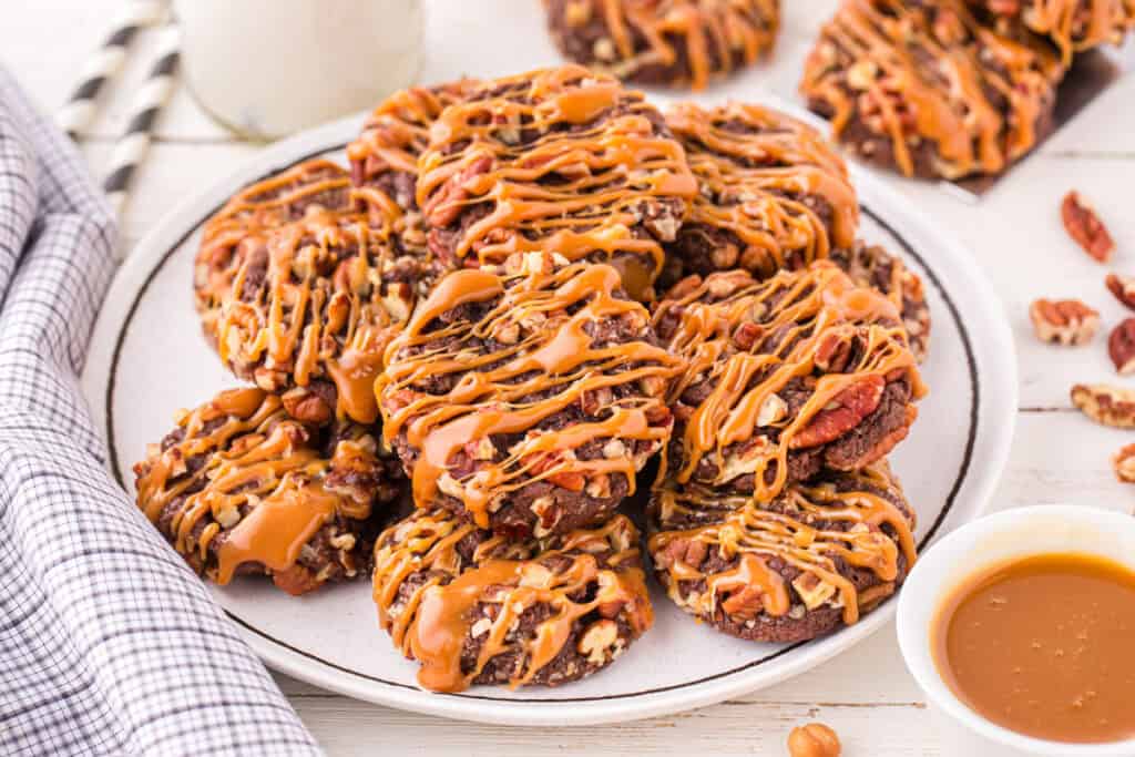 Easy Turtle Cookies piled on a white plate and drizzled with caramel, featuring chocolate cookies coated in chopped pecans and served with a bowl of caramel sauce.