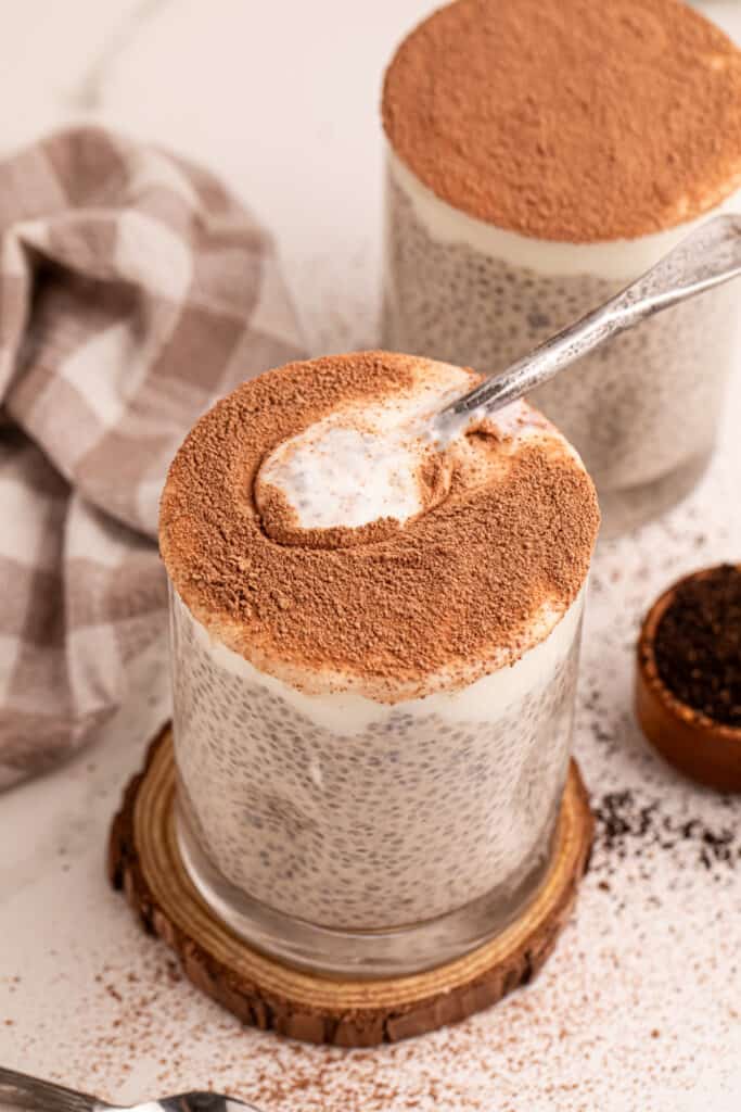 Spoon scooping creamy Tiramisu Chia Pudding from a glass topped with cocoa powder
