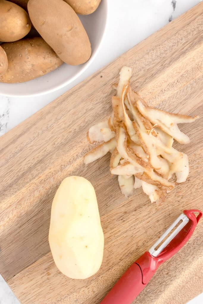 Peeled russet potato on a cutting board with potato peels and a vegetable peeler
