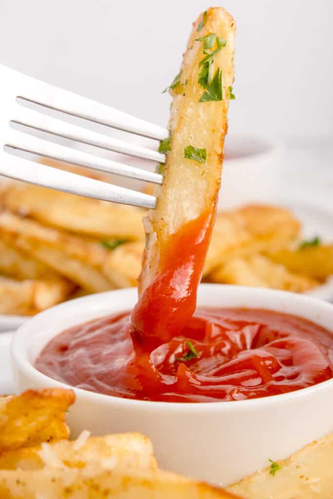Crispy French fry on a fork dipped in ketchup with homemade Air Fryer French Fries in the background