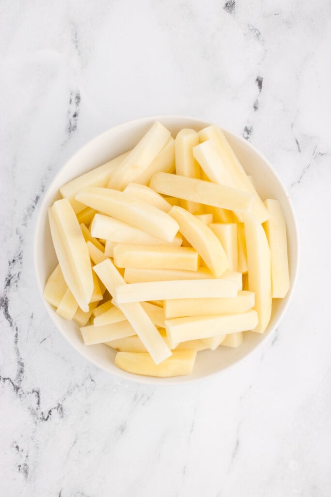 Peeled russet potatoes cut into thick French fry sticks in a white bowl