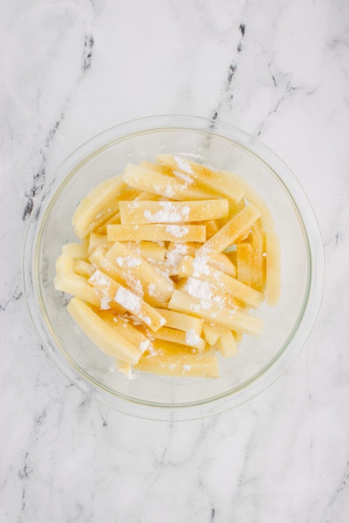 Dried potato fries sprinkled with cornstarch and seasonings in a glass mixing bowl