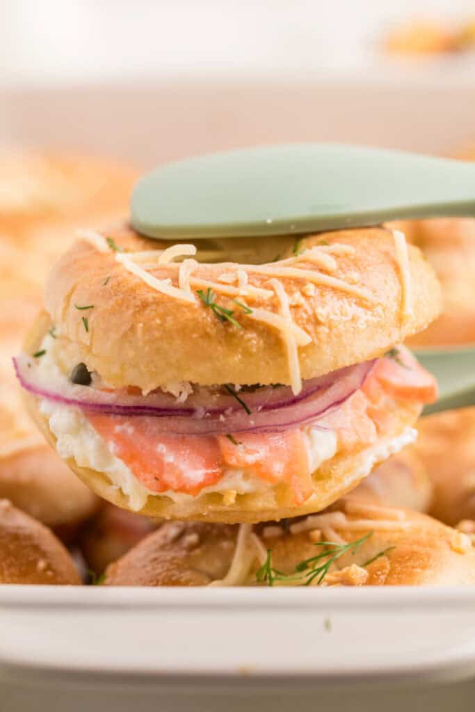 Baked Bagels & Lox Slider lifted from the pan showing smoked salmon, cream cheese, red onion, capers, and dill