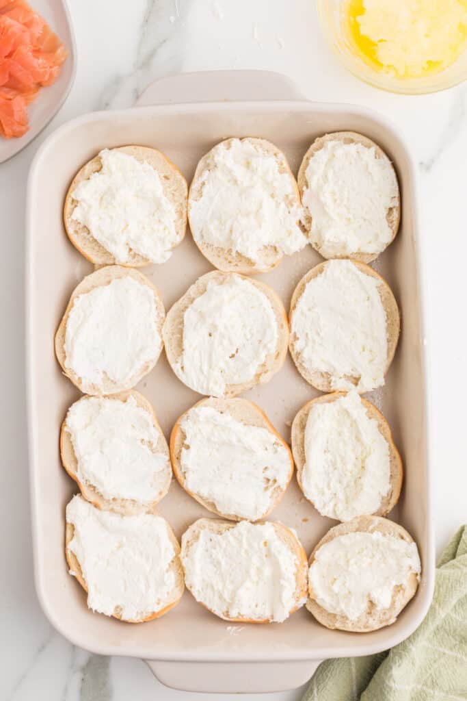 Mini bagel bottoms spread generously with whipped cream cheese in a baking dish