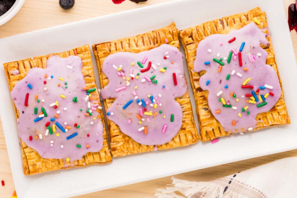Three glazed blueberry pastries with colorful sprinkles arranged on a white serving platter