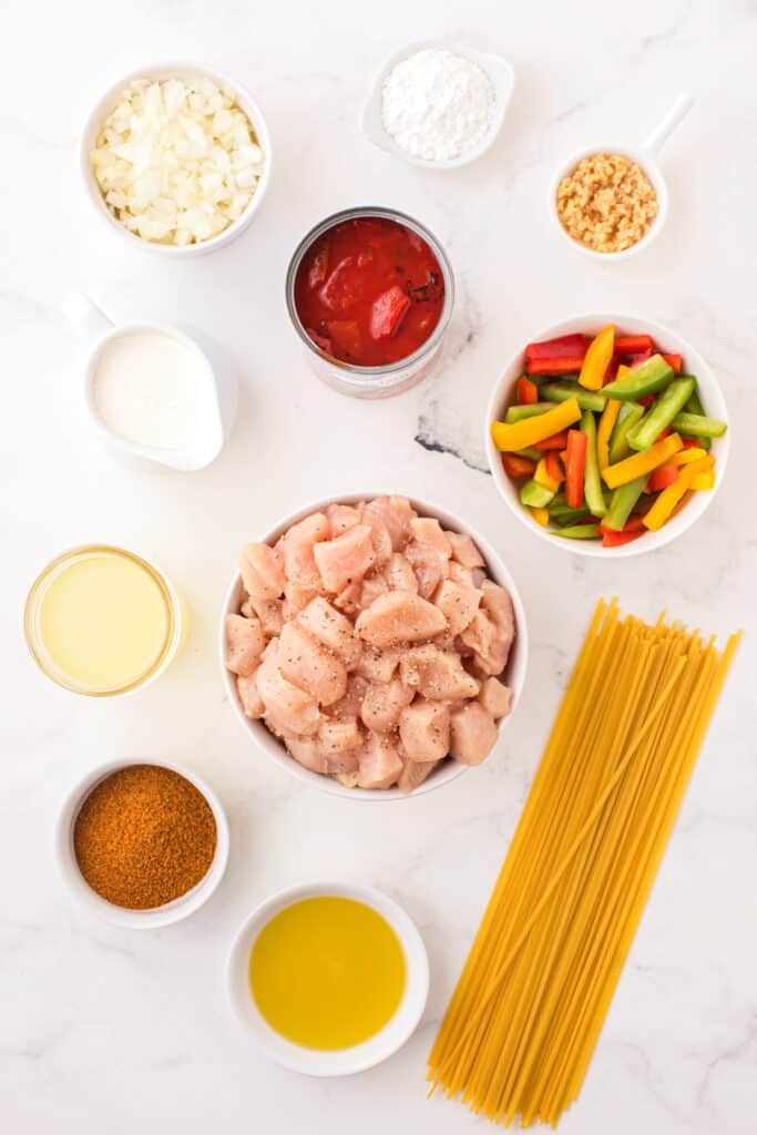 Ingredients for creamy Cajun Chicken Pasta arranged with chicken, linguine, bell peppers, tomatoes, cream, onion, garlic, and seasonings