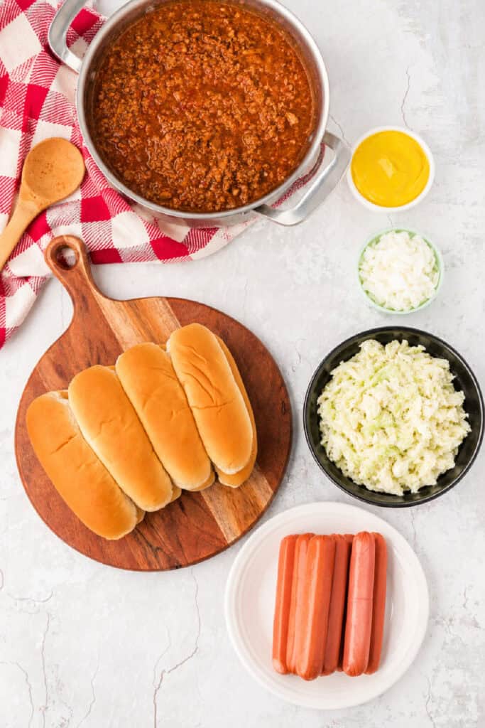 Ingredients for Carolina Dogs including hot dogs, buns, homemade hot dog chili, Southern-style slaw, mustard, and chopped onions arranged for assembly.