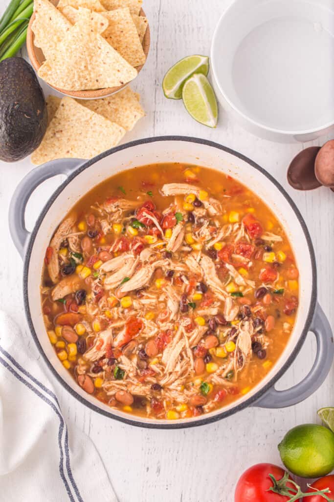 Finished Chicken Taco Soup in a pot with tortilla chips, avocado, lime, and tomatoes nearby.