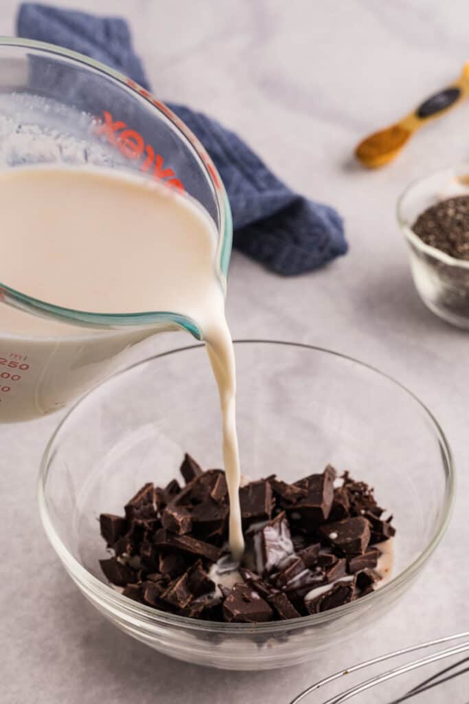 Warm cream mixture being poured over chopped dark chocolate in a glass bowl