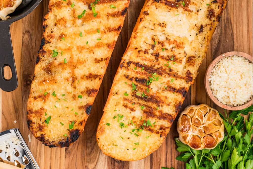 Overhead view of Grilled Garlic Bread with roasted garlic, Parmesan cheese, and fresh parsley on a wooden board.