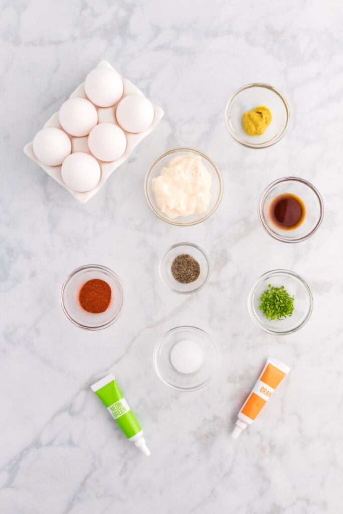 Ingredients for homemade Halloween deviled eggs arranged on a light surface with eggs, mayonnaise, mustard, seasonings, chives, and food coloring.