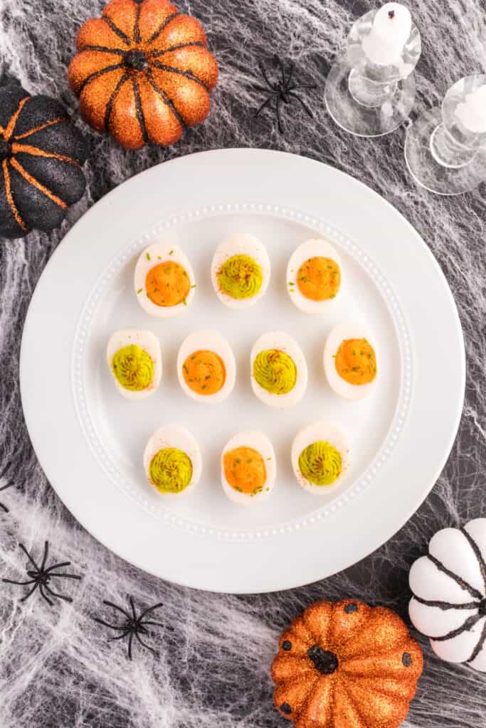 Halloween deviled eggs with orange and green filling served on a white platter surrounded by spooky pumpkins, spiders, and cobwebs.