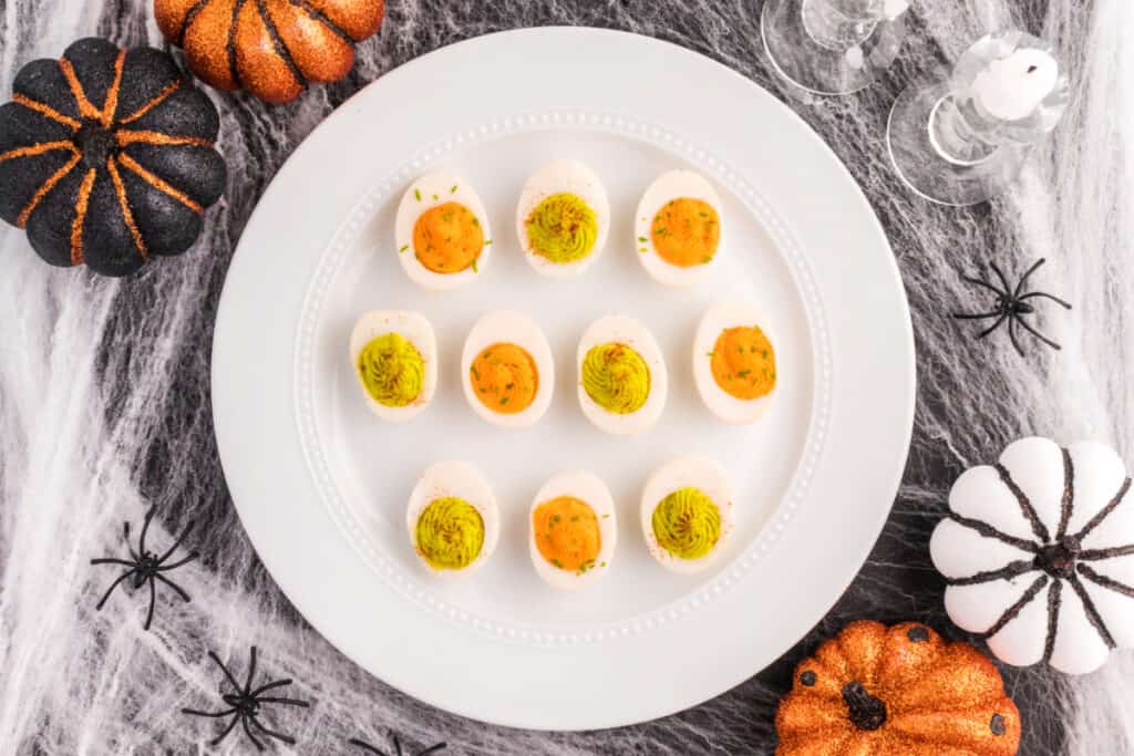 Overhead view of colorful Halloween deviled eggs surrounded by black and orange pumpkins, spiders, candles, and cobweb decorations.