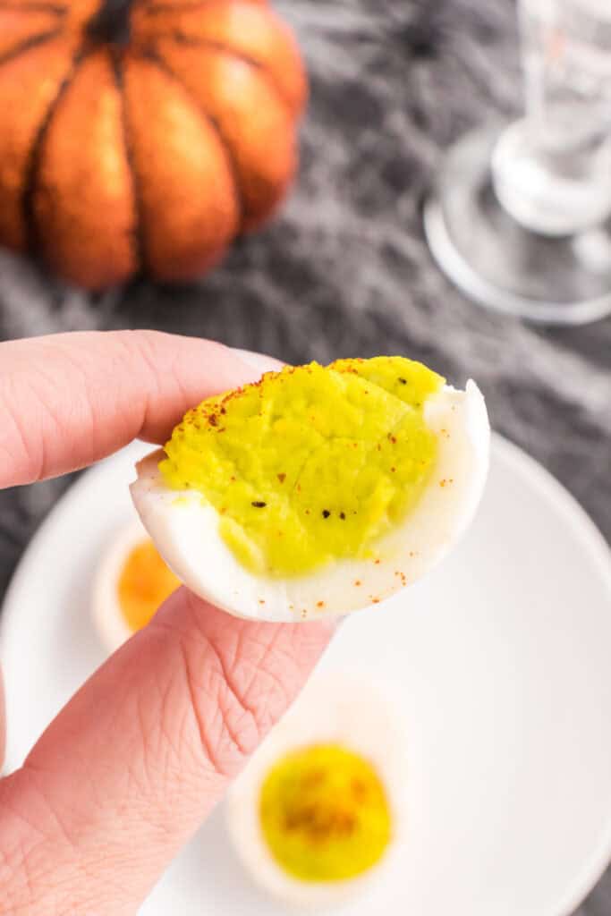 Hand holding a neon green Halloween deviled egg sprinkled with paprika over a serving plate.