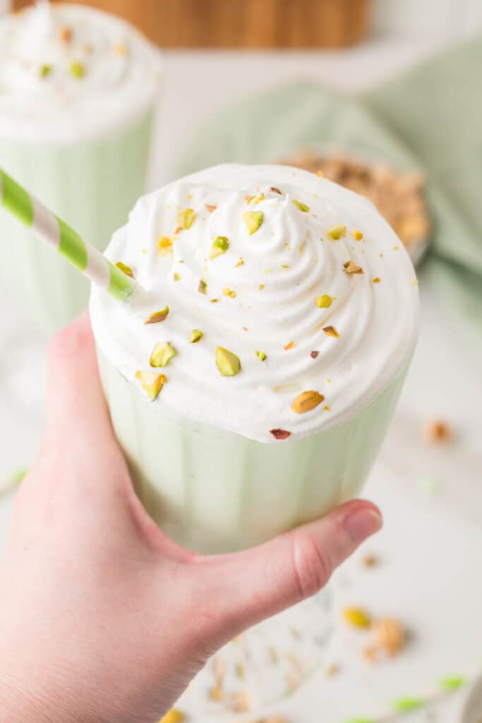 Hand holding a pistachio milkshake topped with whipped cream, chopped pistachios, and a striped straw