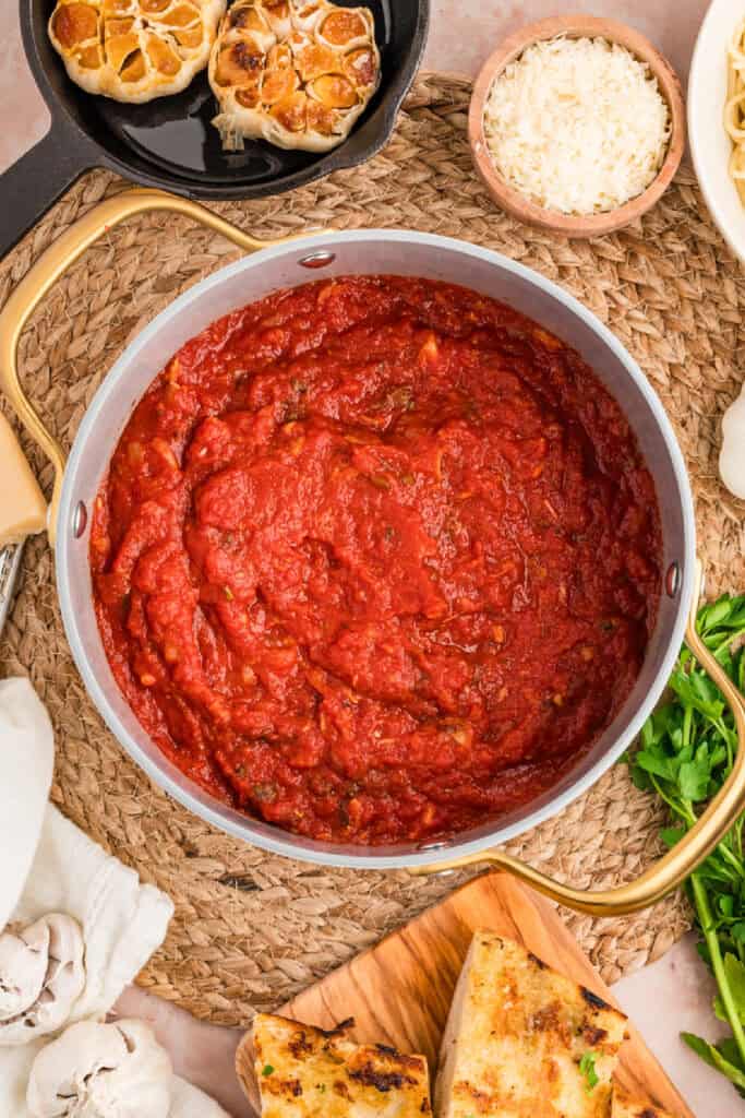 Finished homemade roasted garlic tomato sauce in a saucepan with roasted garlic and Parmesan nearby.