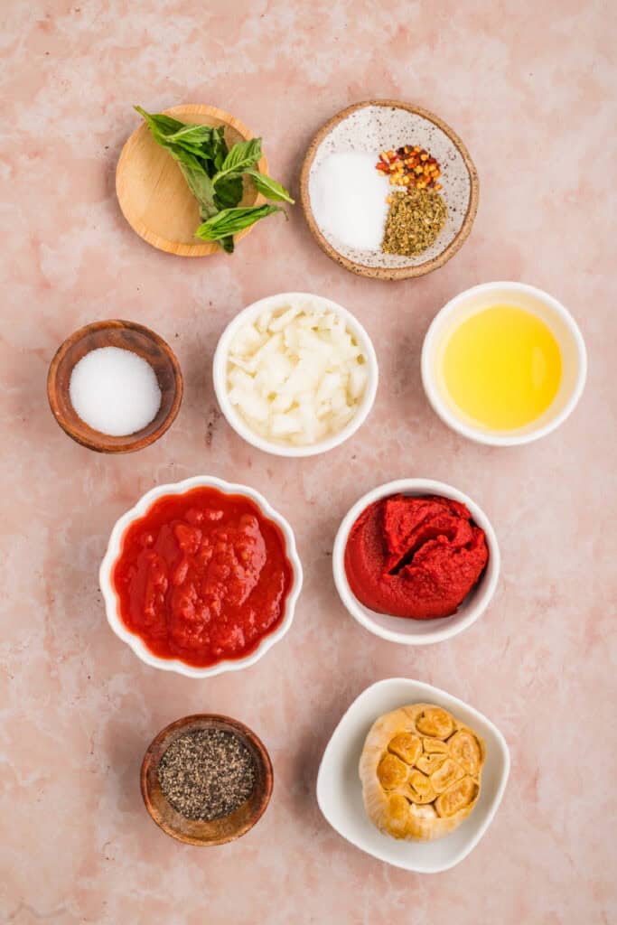 Ingredients for homemade roasted garlic pasta sauce arranged on a pink surface, including roasted garlic, tomatoes, onion, basil, and seasonings.