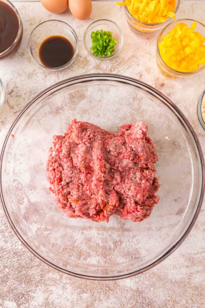 Ground beef and pork in a mixing bowl surrounded by eggs, peppers, cheese, and sauces