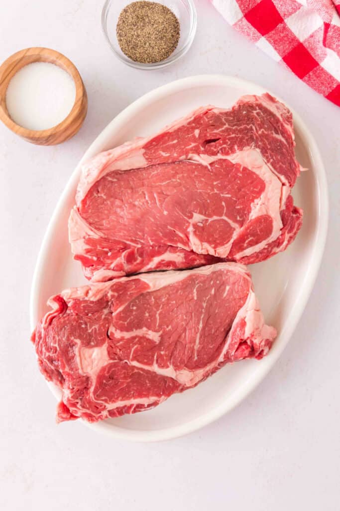 Raw ribeye steaks on a white platter with salt and ground black pepper ready for seasoning.