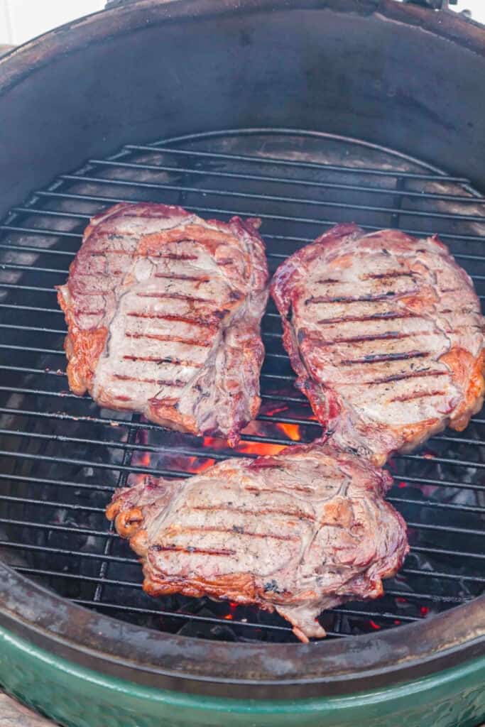 Three ribeye steaks searing over hot coals with dark grill marks forming on the surface.