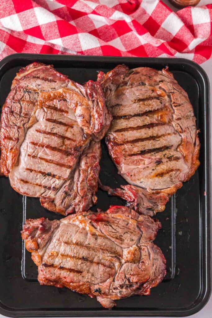 Three finished ribeye steaks with browned edges and grill marks resting on a tray.