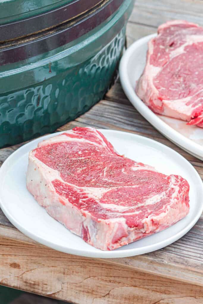 Thick ribeye steak seasoned with salt and pepper beside a BBQ grill.