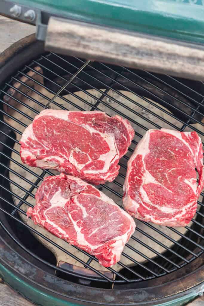 Three seasoned ribeye steaks cooking over indirect heat on a BBQ grill.