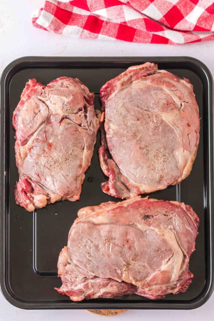 Three partially cooked ribeye steaks resting on a tray before searing.