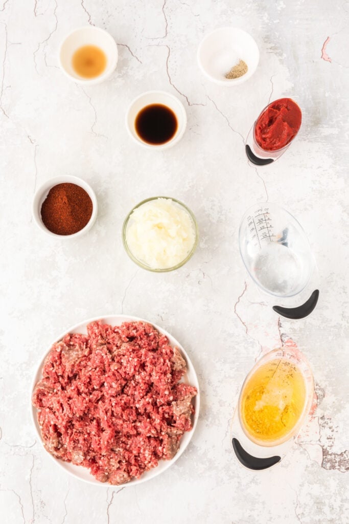 Ingredients for Southern Style Hot Dog Chili including ground beef, chopped onion, tomato paste, beer, Worcestershire sauce, chili powder, vinegar, salt, and pepper arranged on a light background.