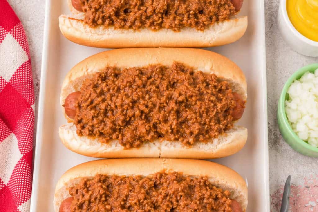 Three hot dogs topped with homemade Southern Style Hot Dog Chili on a white serving platter with diced onions and mustard nearby.