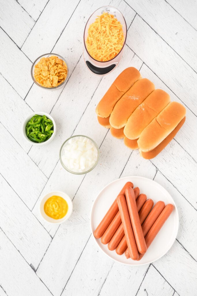 Ingredients for Texas Chili Dogs including hot dogs, buns, cheddar cheese, onions, jalapeños, mustard, and corn chips