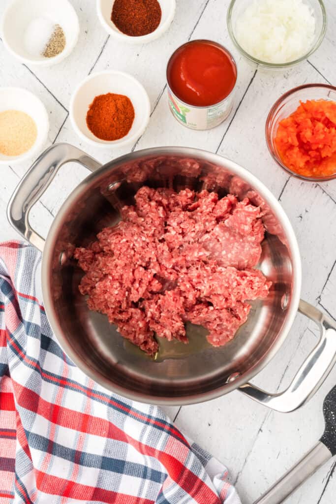 Ground beef in a large pot with tomato sauce, peppers, onion, and seasonings nearby