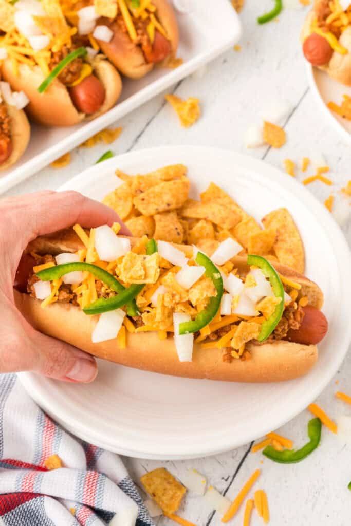 Hand holding a loaded Texas Chili Dog topped with chili, cheese, onions, jalapeños, and corn chips