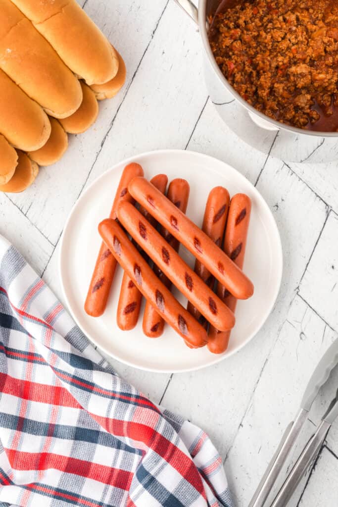 Grilled hot dogs on a white plate with buns and homemade chili nearby