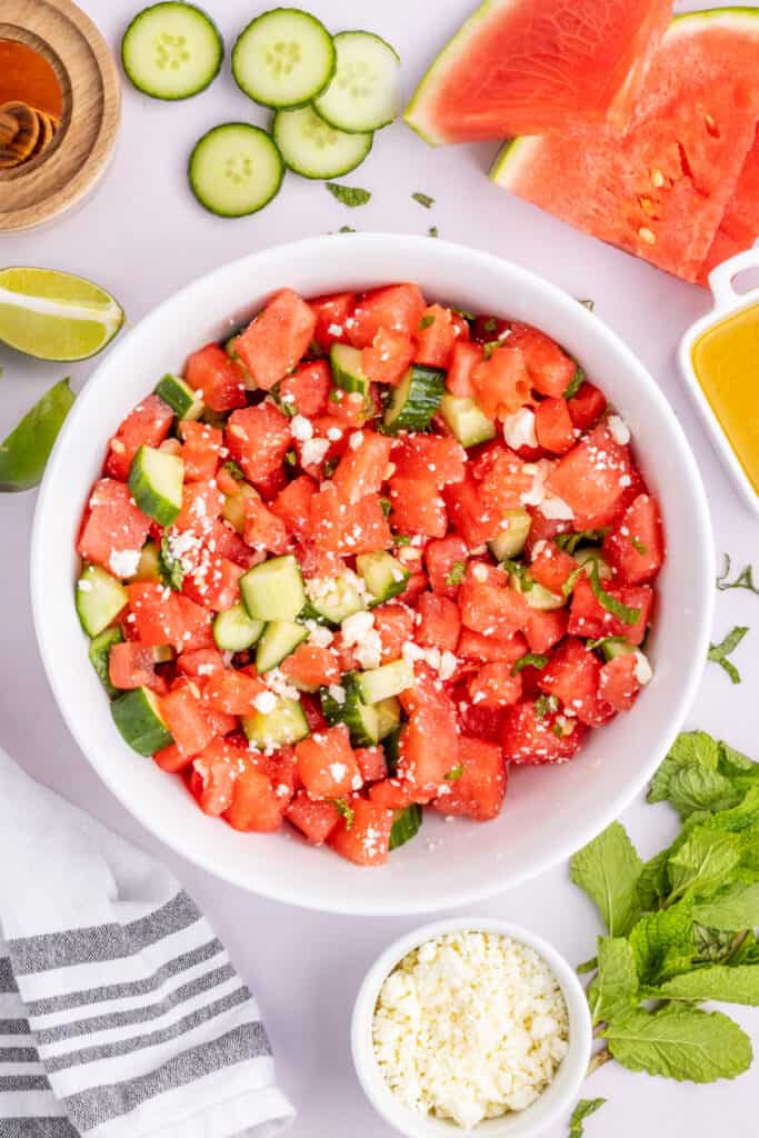 Watermelon Feta Mint Salad served in a large white bowl surrounded by fresh watermelon, cucumber, mint, and feta.