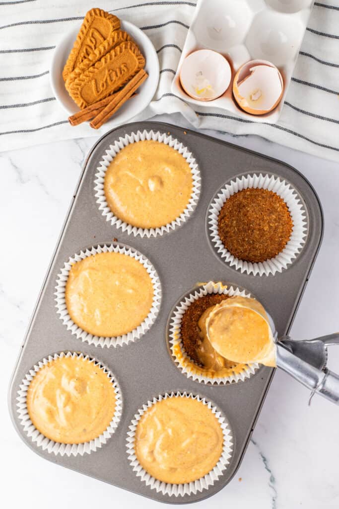 Pumpkin cheesecake batter being portioned over Biscoff cookie crusts in a lined muffin pan.