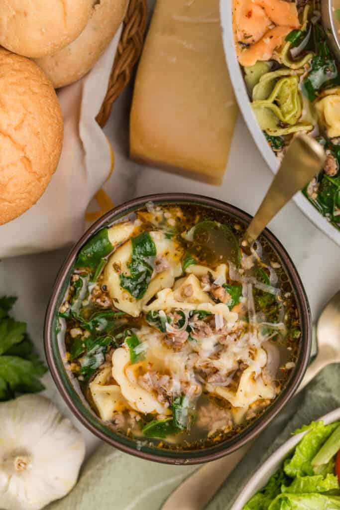 Bowl of sausage tortellini soup topped with shredded Parmesan and surrounded by bread, garlic, and fresh greens.