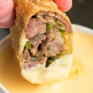 Philly Cheesesteak Egg Roll dipped in cheese sauce with steak, peppers, onions, and melted provolone visible inside