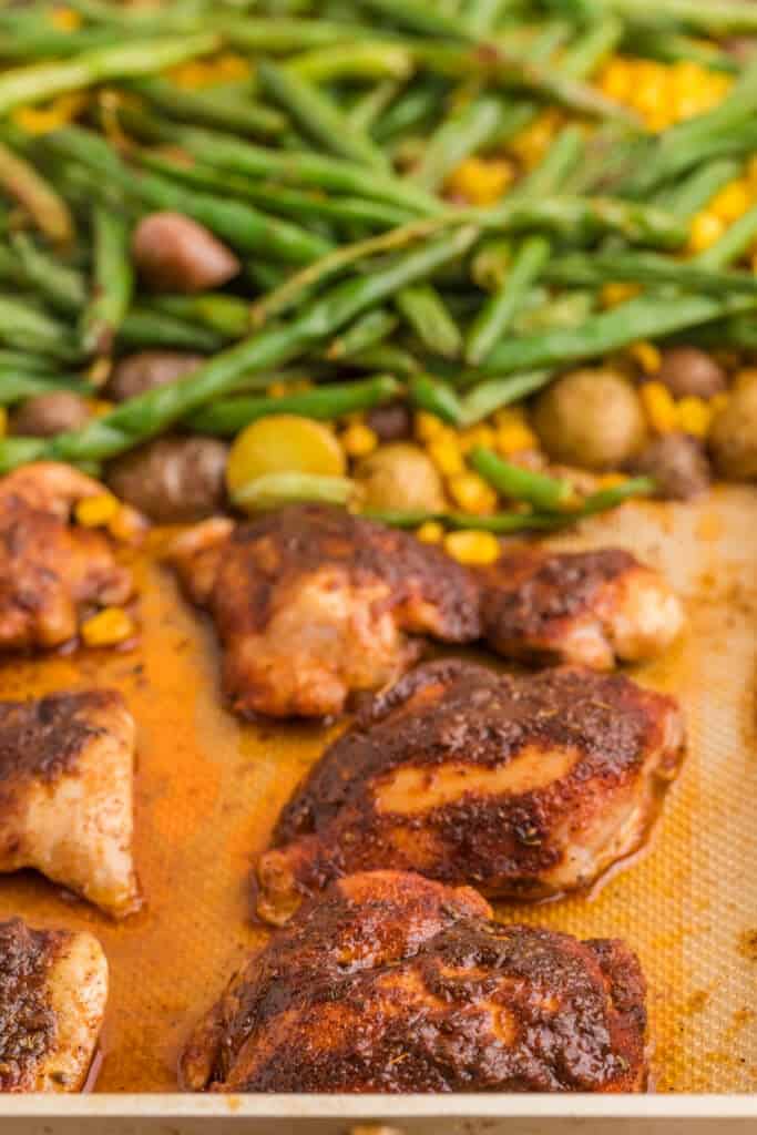Close-up of roasted blackened chicken thighs with green beans, corn, and baby potatoes on the sheet pan.