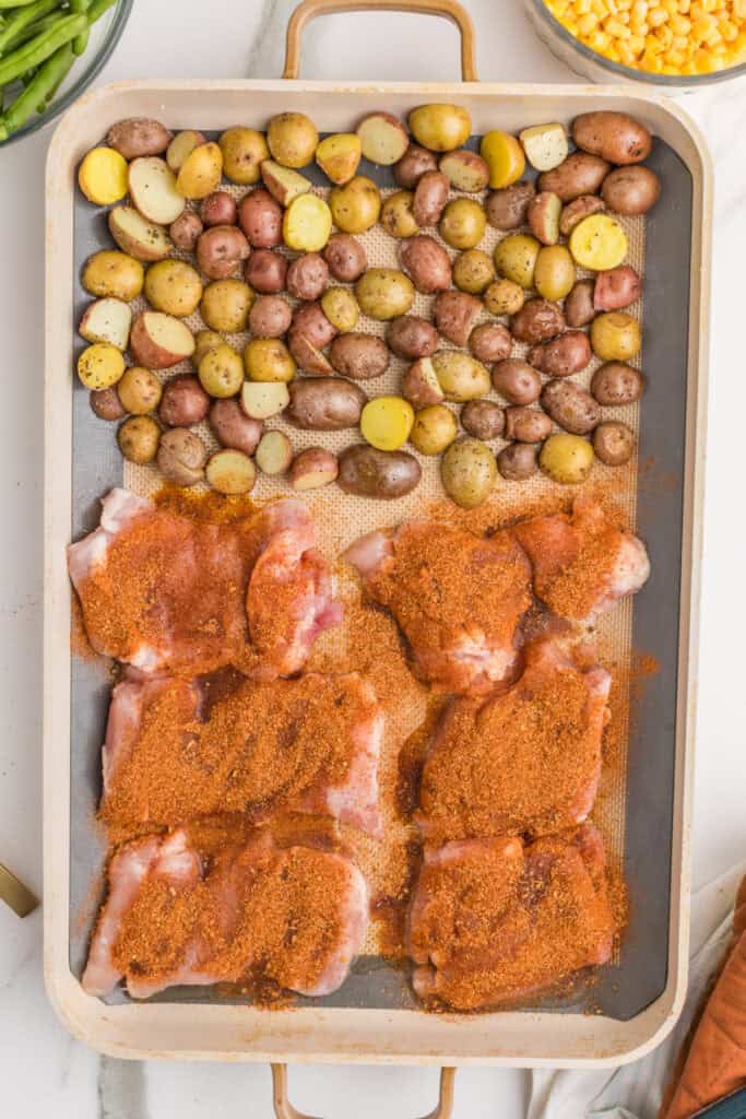Boneless chicken thighs coated with blackened seasoning beside partially roasted baby potatoes on a sheet pan.