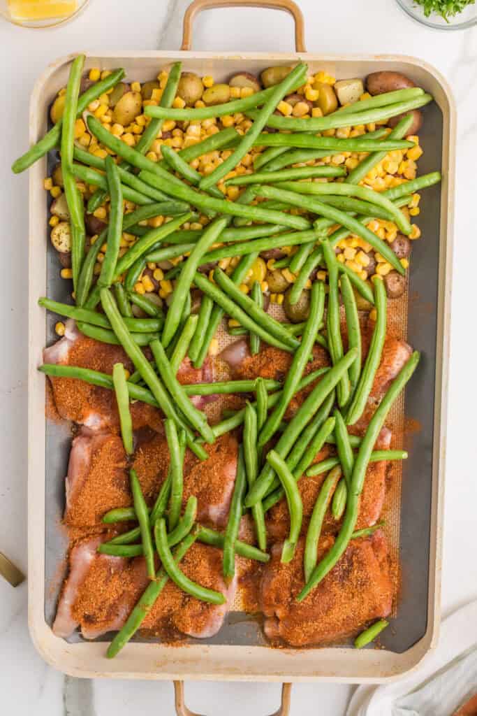 Blackened chicken thighs, baby potatoes, green beans, and corn arranged on a sheet pan before roasting.
