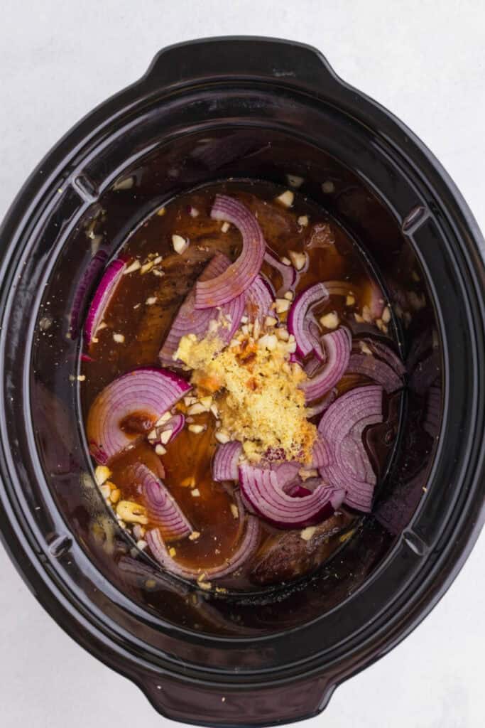 Seared chuck roast in a slow cooker with sliced onion, fresh ginger, garlic, beef stock, and soy sauce