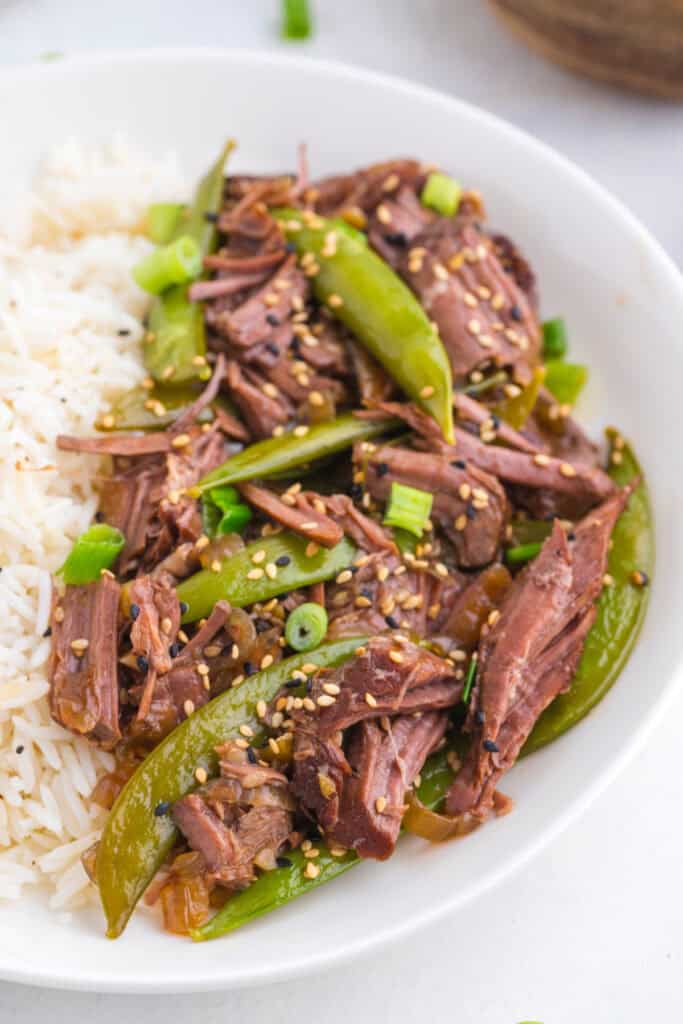 Close-up of tender shredded Asian pot roast with snap peas, green onions, sesame seeds, and white rice