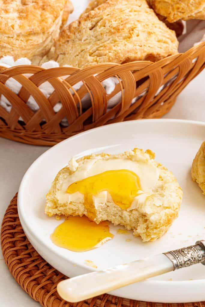 Split sour cream biscuit topped with butter and honey on a white plate with more biscuits in a basket.