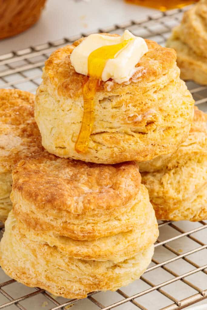 Flaky air fryer sour cream biscuits on a cooling rack, with one biscuit topped with butter and dripping honey.
